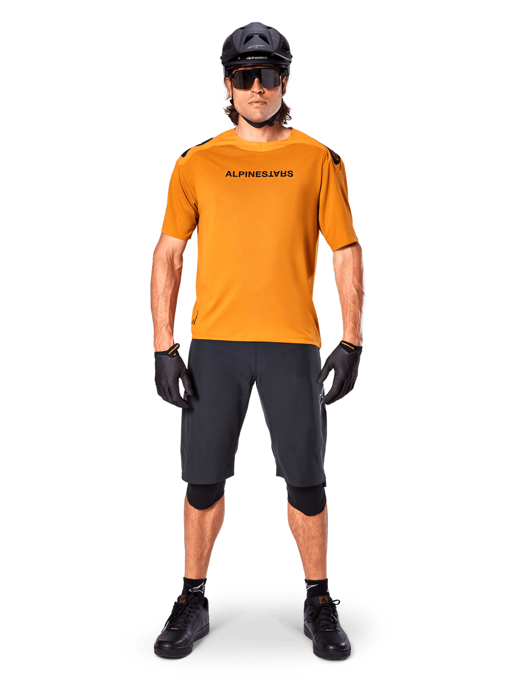 Ciclista masculino vistiendo el jersey de manga corta Alpinestars A-Aria Polartec Switch en color Dark Gold, combinado con pantalones cortos de MTB negros, casco, gafas de sol y guantes, equipamiento profesional para ciclismo de montaña