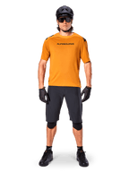 Ciclista masculino vistiendo el jersey de manga corta Alpinestars A-Aria Polartec Switch en color Dark Gold, combinado con pantalones cortos de MTB negros, casco, gafas de sol y guantes, equipamiento profesional para ciclismo de montaña