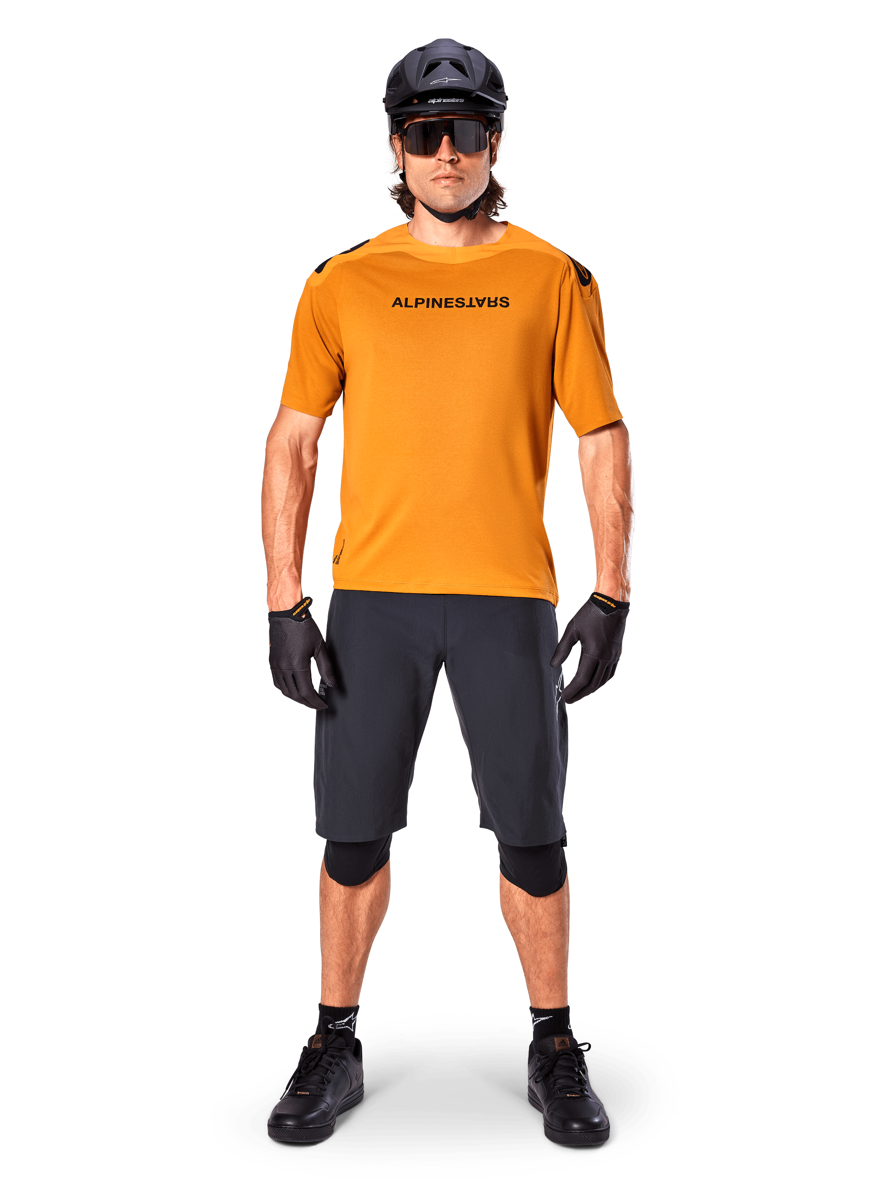 Ciclista masculino vistiendo el jersey de manga corta Alpinestars A-Aria Polartec Switch en color Dark Gold, combinado con pantalones cortos de MTB negros, casco, gafas de sol y guantes, equipamiento profesional para ciclismo de montaña