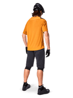 Alpinestars A-Aria Polartec Switch Jersey, jersey de MTB de manga corta, Dark Gold, vista trasera del modelo con pantalones cortos de montaña gris oscuro, casco negro y guantes, ropa técnica de ciclismo para senderos