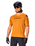 Jersey Alpinestars A-Aria Polartec Switch, jersey de MTB de manga corta, color oro oscuro con logotipos en negro, usado por un modelo con casco negro, gafas de sol y guantes de ciclismo, ropa de ciclismo de montaña de alto rendimiento