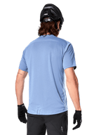 Alpinestars A-Dura Dri Astar Short Sleeve Jersey, jersey de MTB, color Azul Infinity, vista trasera que muestra paneles de ventilación de malla en los hombros y una parte trasera más larga, usado con casco de MTB y guantes negros, equipamiento de ciclismo de alto rendimiento