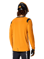 Jersey de manga larga Alpinestars A-Dura Dri Wool en color Dark Gold, vista posterior de un ciclista con casco de mountain bike y guantes, con paneles de malla negros en los hombros y una parte trasera más larga, ropa técnica de ciclismo para trail