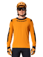 Modelo masculino con jersey Alpinestars A-Dura Dri Wool de manga larga en color Dark Gold, coordinado con casco de MTB y guantes dorados a juego, paneles de malla negros en hombros y mangas, logotipo Alpinestars en el pecho, gafas de sol y pantalones oscuros, equipamiento técnico para bicicleta de montaña