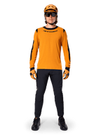 Ciclista de montaña con el jersey de manga larga Alpinestars A-Dura Dri Wool, color oro oscuro con detalles negros en el cuello y las mangas, combinado con pantalones negros, casco y guantes dorados a juego y gafas de sol negras, equipamiento profesional para ciclismo de montaña