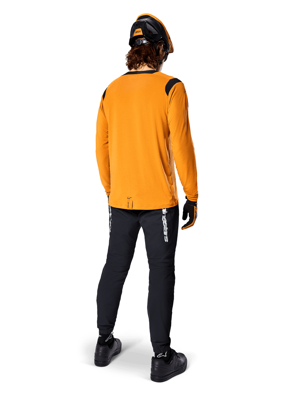 Ciclista de montaña visto desde atrás con la camiseta de manga larga Alpinestars A-Dura Dri Wool en color Dark Gold, pantalones técnicos negros con logotipos blancos y un casco a juego en negro y naranja, ropa de ciclismo de montaña de alto rendimiento.