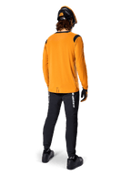 Ciclista de montaña visto desde atrás con la camiseta de manga larga Alpinestars A-Dura Dri Wool en color Dark Gold, pantalones técnicos negros con logotipos blancos y un casco a juego en negro y naranja, ropa de ciclismo de montaña de alto rendimiento.