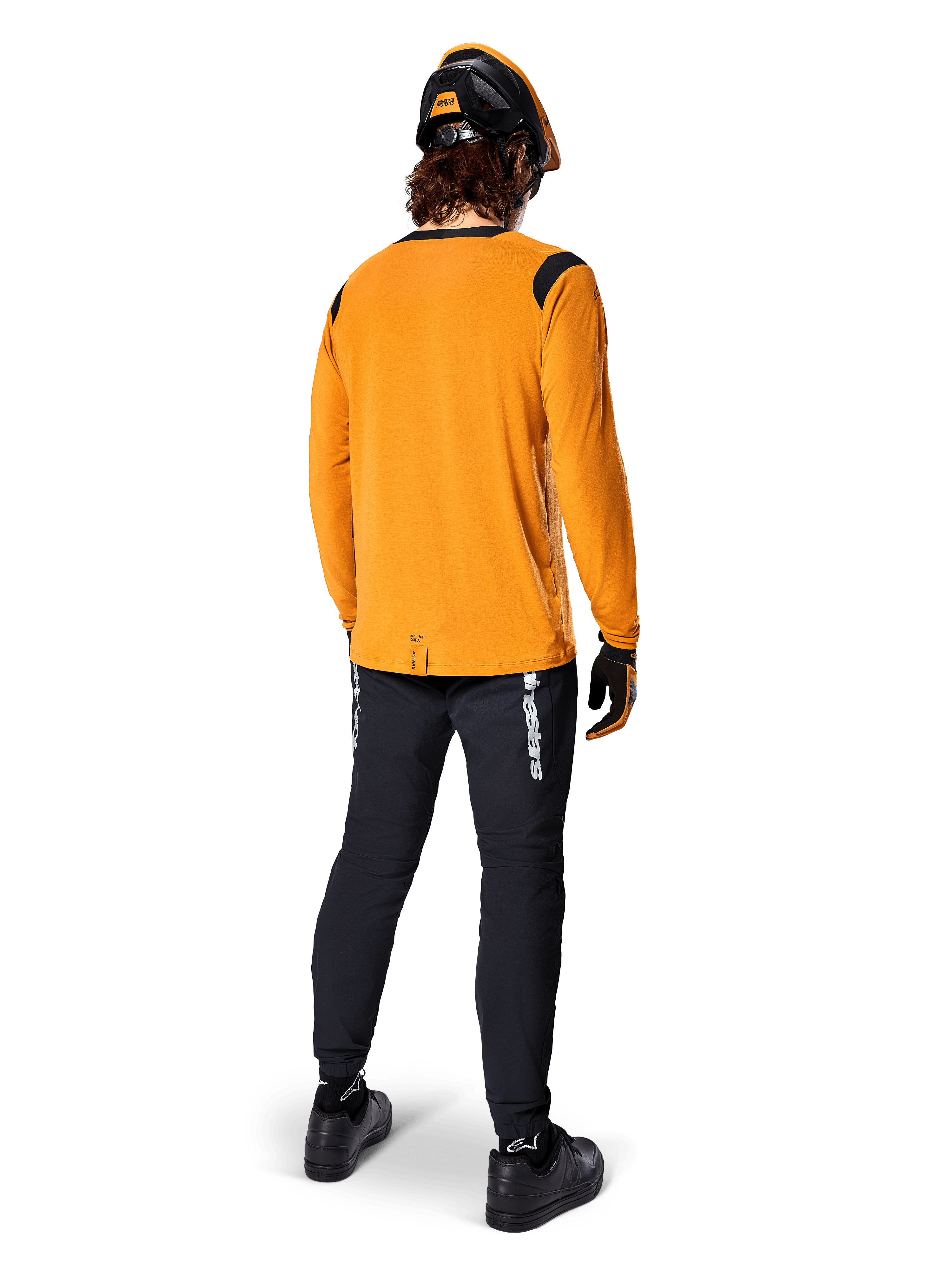 Ciclista de montaña visto desde atrás con la camiseta de manga larga Alpinestars A-Dura Dri Wool en color Dark Gold, pantalones técnicos negros con logotipos blancos y un casco a juego en negro y naranja, ropa de ciclismo de montaña de alto rendimiento.
