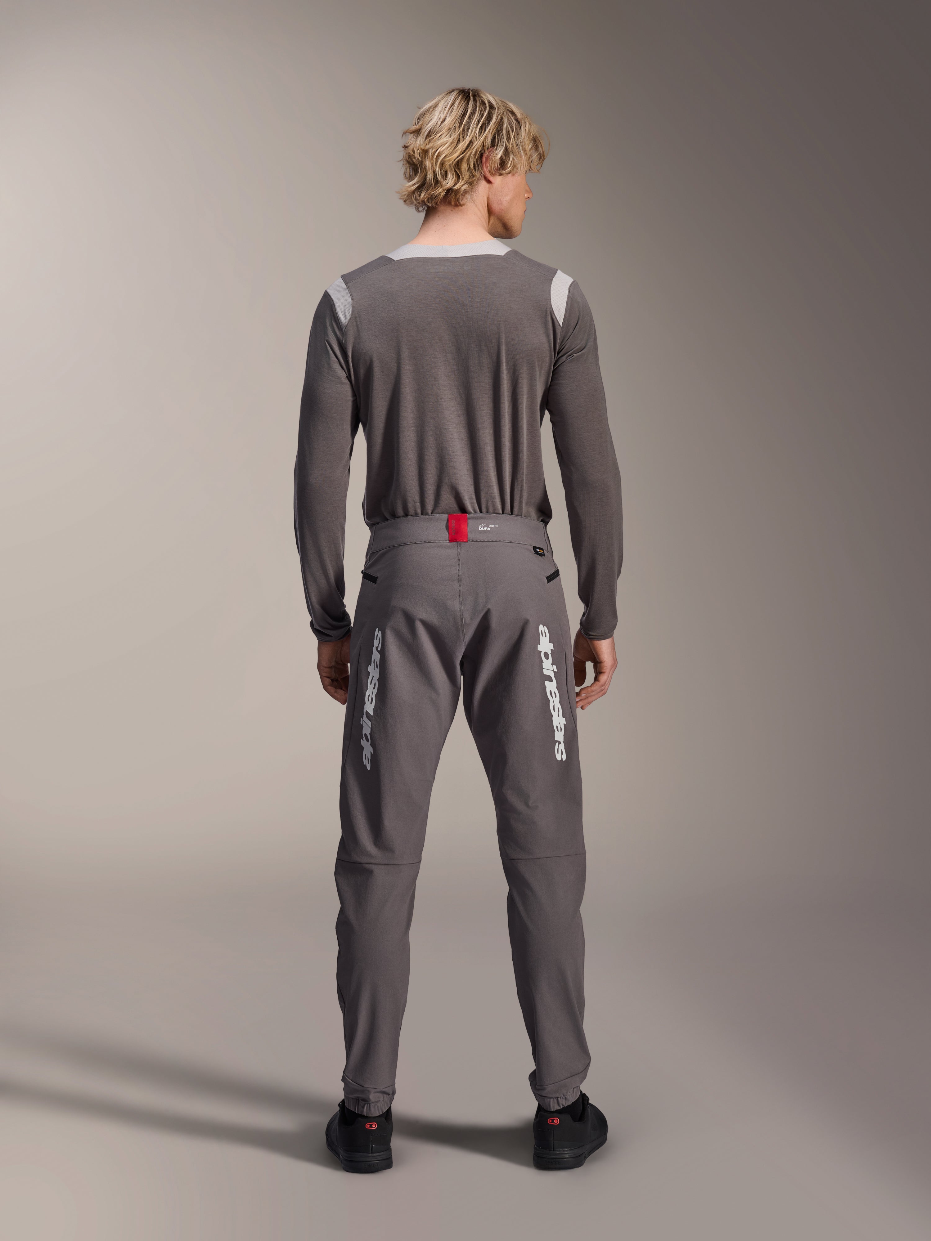 Modelo de espaldas vistiendo pantalones Alpinestars A-Dura Elite en gris oscuro y un jersey de manga larga gris a juego, con logotipos blancos de Alpinestars en las piernas, un detalle rojo en la cintura y zapatillas negras, equipamiento profesional para ciclismo MTB.