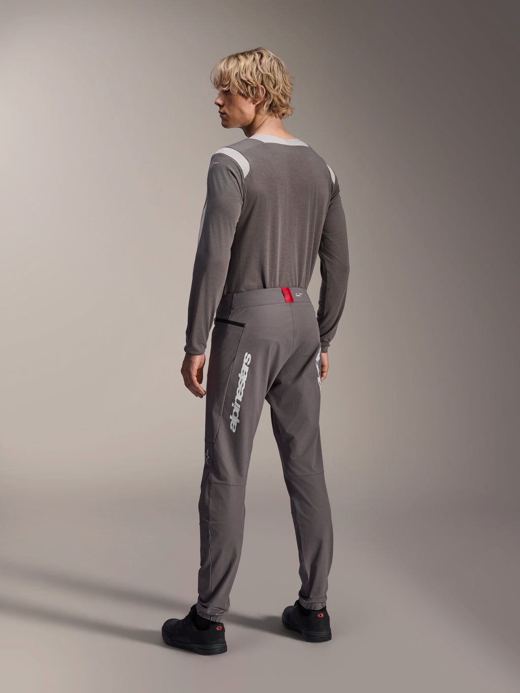 Modelo desde una vista posterior con equipamiento MTB de Alpinestars, jersey de manga larga A-Dura Dri Wool gris, pantalones A-Dura Elite gris oscuro con marca vertical blanca y detalle rojo en la cintura, conjunto de ciclismo coordinado para mountain bike