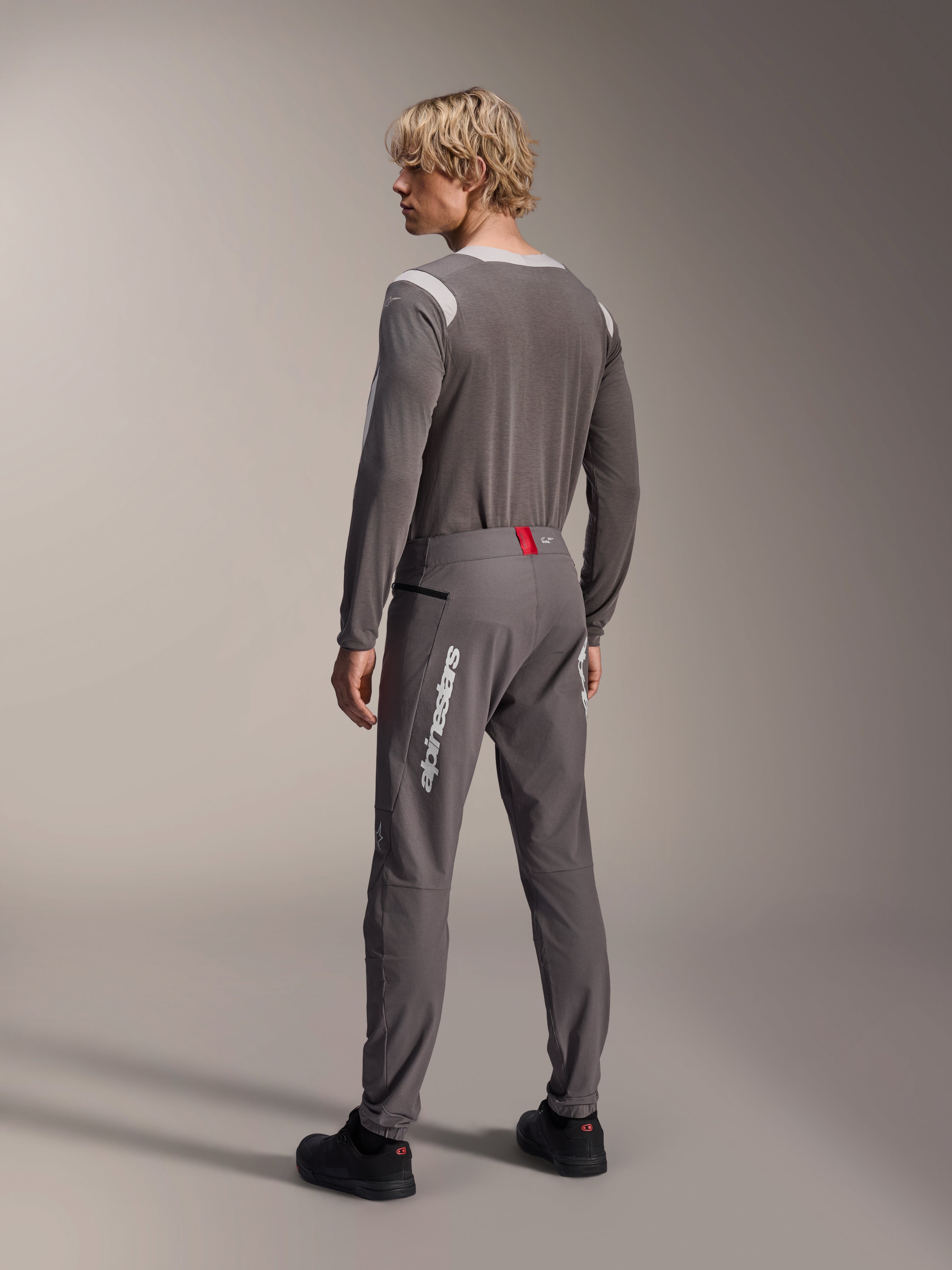 Modelo desde una vista posterior con equipamiento MTB de Alpinestars, jersey de manga larga A-Dura Dri Wool gris, pantalones A-Dura Elite gris oscuro con marca vertical blanca y detalle rojo en la cintura, conjunto de ciclismo coordinado para mountain bike