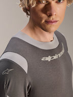 Alpinestars A-Dura Dri Wool Jersey - Manga Larga, jersey de ciclismo de montaña gris oscuro, primer plano de modelo masculino que muestra el cuello unido elástico y los paneles de malla en los hombros, con el logotipo de Alpinestars en el pecho y la manga