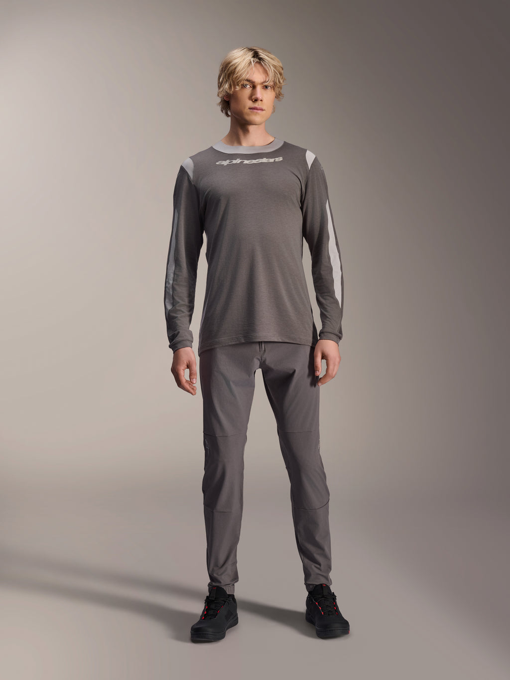 Jersey Alpinestars A-Dura Dri Wool de manga larga, gris oscuro, usado por un modelo con pantalones grises a juego y calzado negro, logotipo de Alpinestars en blanco en el pecho, paneles de malla gris claro en hombros y mangas, equipamiento técnico para bicicleta de montaña