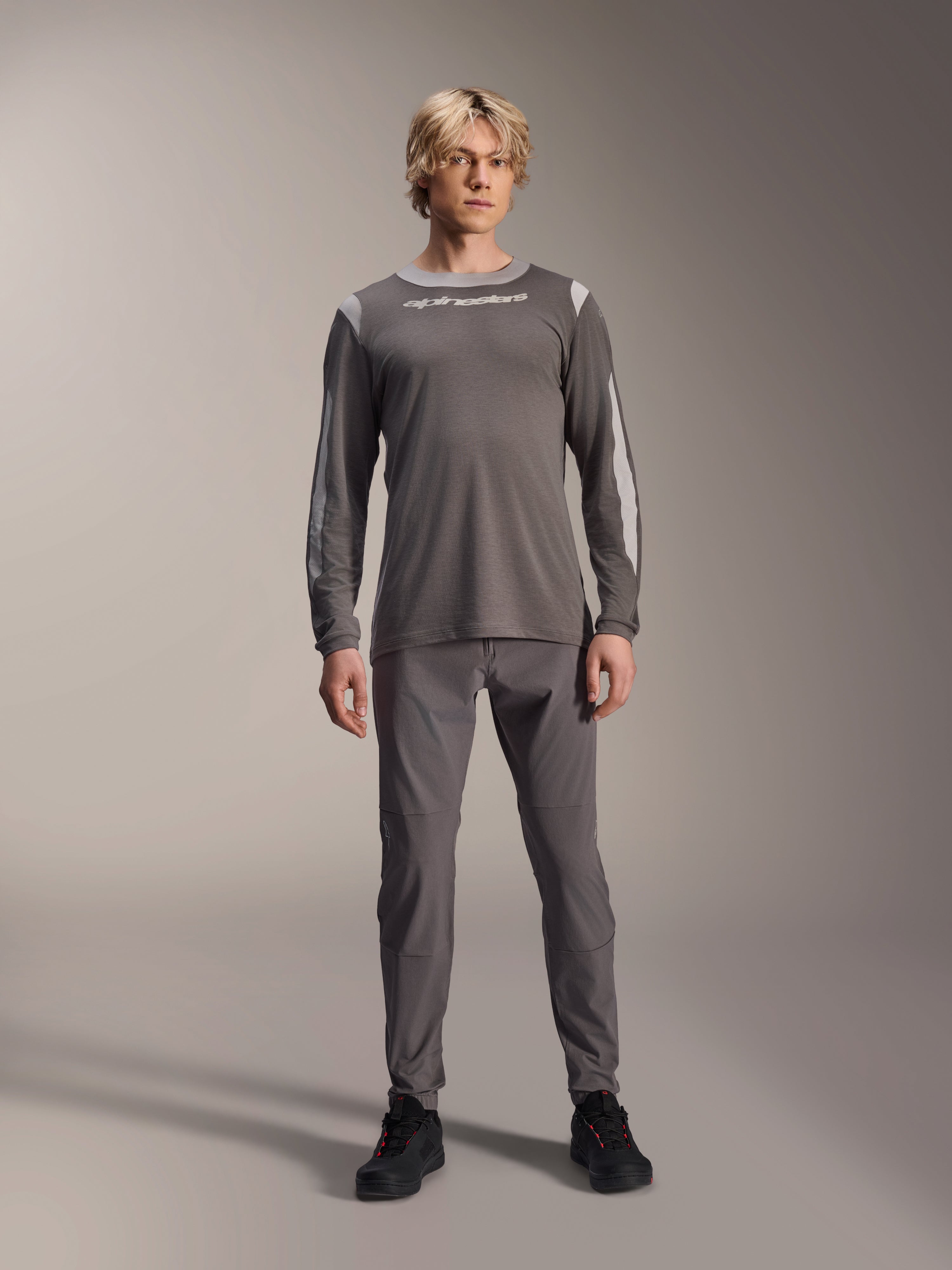 Jersey Alpinestars A-Dura Dri Wool de manga larga, gris oscuro, usado por un modelo con pantalones grises a juego y calzado negro, logotipo de Alpinestars en blanco en el pecho, paneles de malla gris claro en hombros y mangas, equipamiento técnico para bicicleta de montaña
