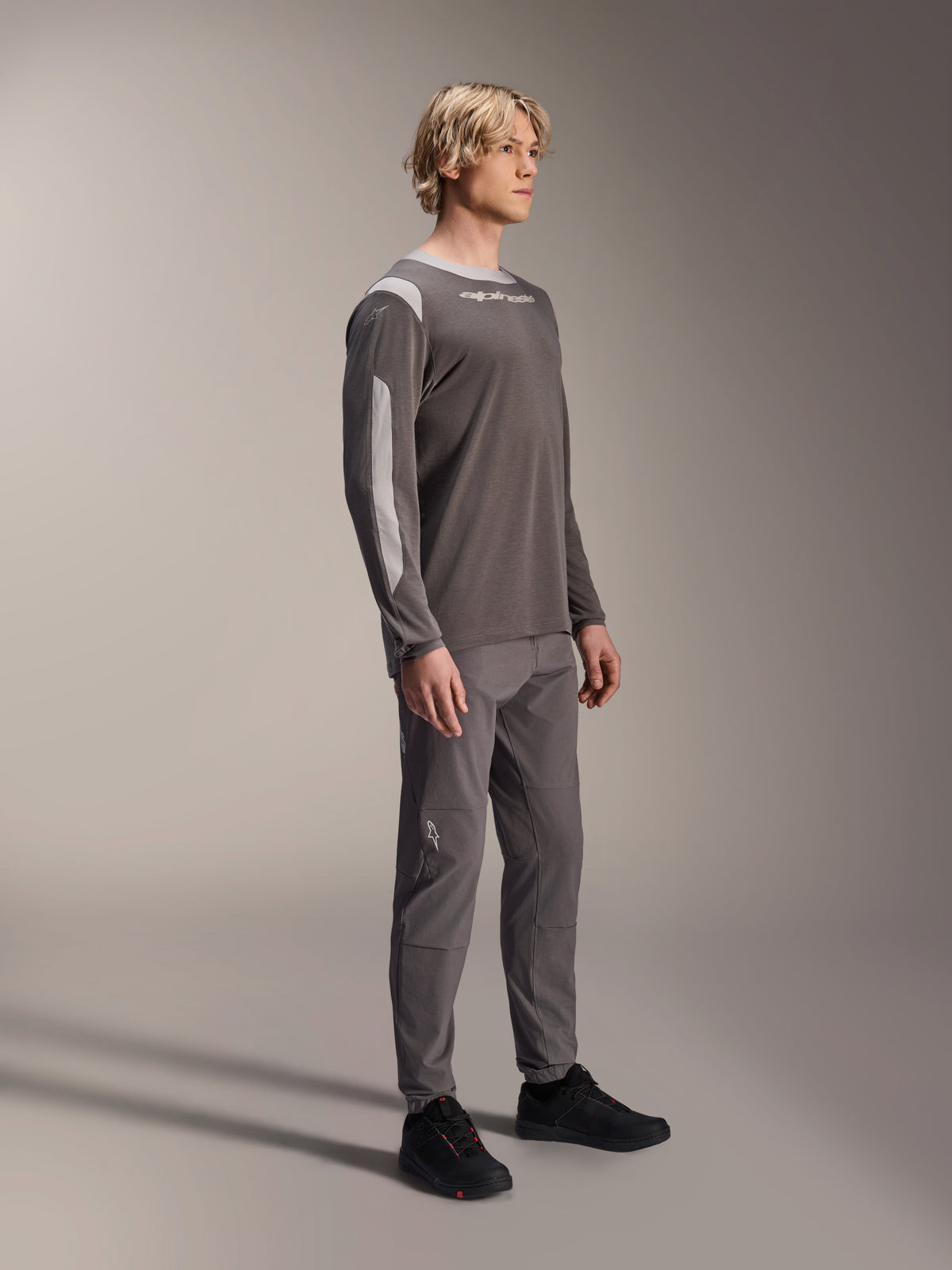 Modelo masculino vistiendo el jersey de manga larga Alpinestars A-Dura Dri Wool en gris oscuro, combinado con pantalones grises a juego y calzado negro, con paneles de malla gris claro en los hombros y las mangas, equipamiento técnico de MTB para ciclismo de montaña