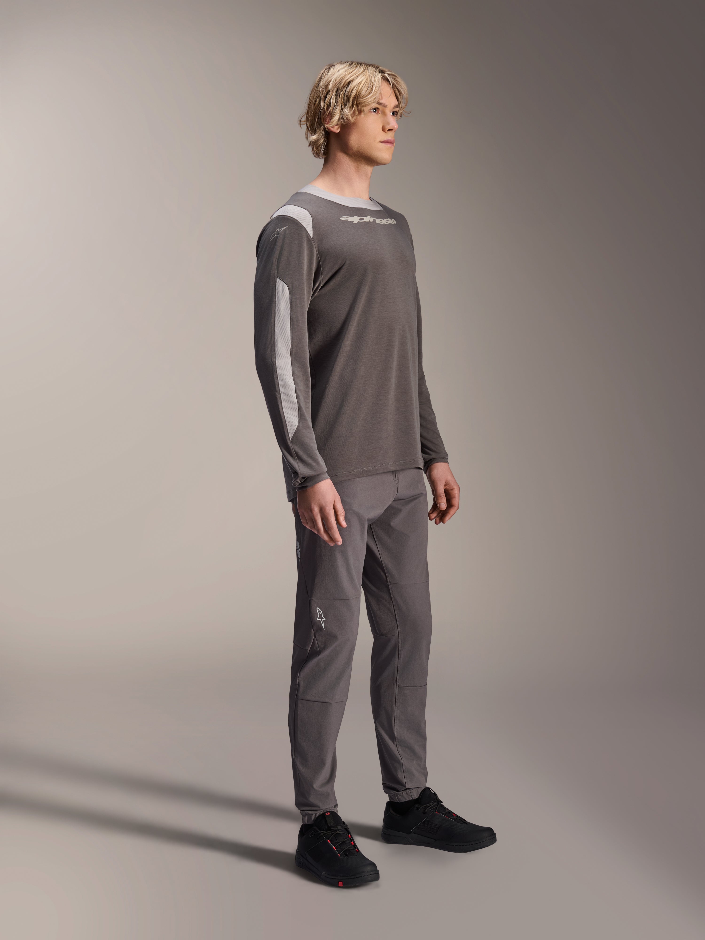 Modelo masculino vistiendo el jersey de manga larga Alpinestars A-Dura Dri Wool en gris oscuro, combinado con pantalones grises a juego y calzado negro, con paneles de malla gris claro en los hombros y las mangas, equipamiento técnico de MTB para ciclismo de montaña