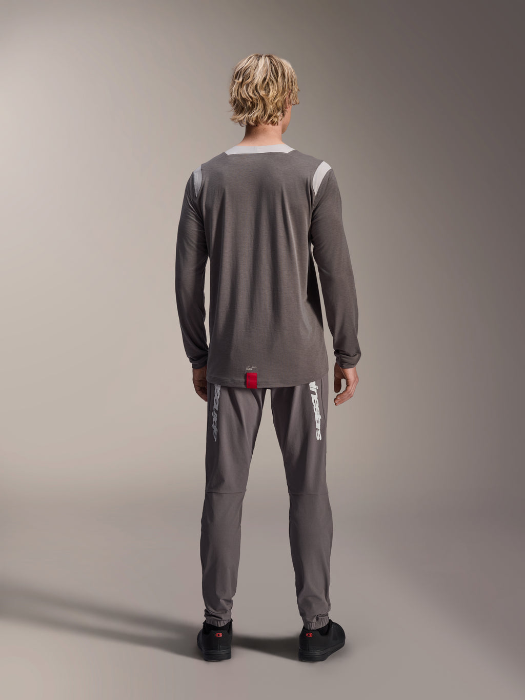 Alpinestars A-Dura Dri Wool Jersey de manga larga, gris oscuro, vista trasera en modelo, con paneles de malla gris claro en los hombros y el cuello, dobladillo trasero más largo con una pequeña etiqueta roja de la marca, combinado con pantalones de ciclismo grises a juego.