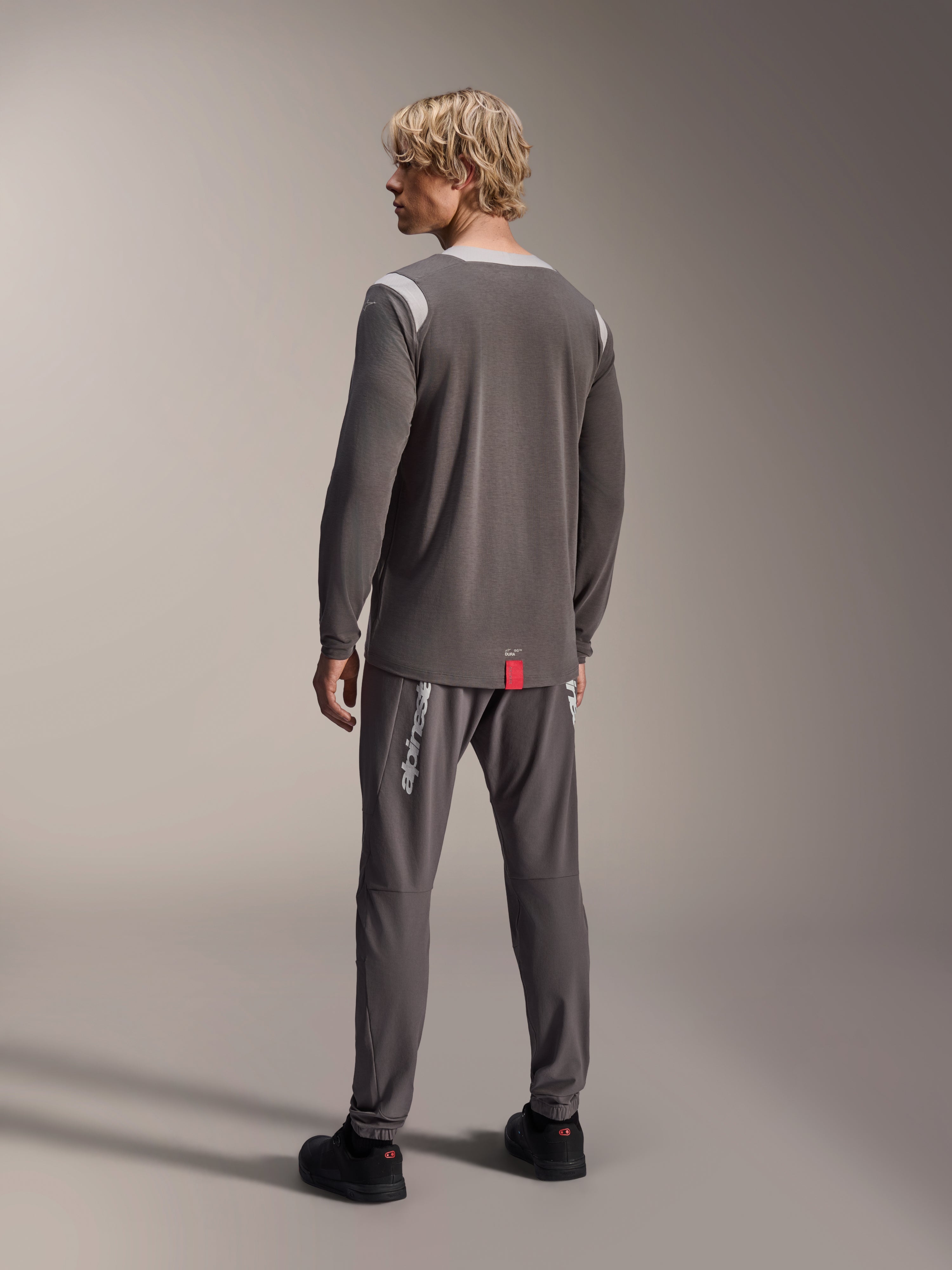 Modelo con jersey Alpinestars A-Dura Dri Wool de manga larga, gris oscuro con paneles de malla en los hombros en gris claro, combinado con pantalones de ciclismo a juego en gris oscuro, vista trasera que muestra el dobladillo trasero más largo, ropa para bicicleta de montaña