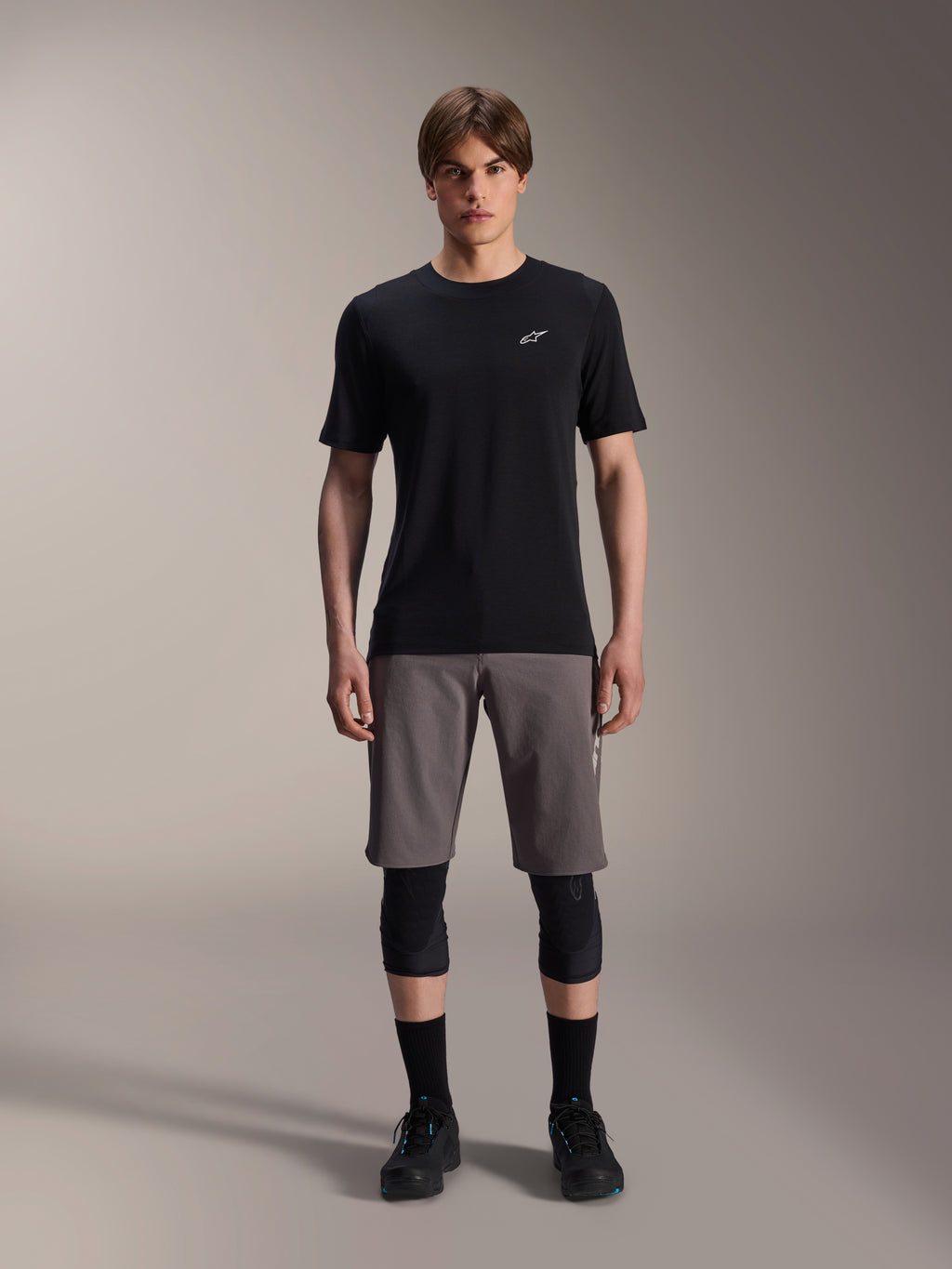 Jersey de manga corta Alpinestars A-Dura Dri Wool, negro, usado por un modelo con pantalones cortos de MTB grises, rodilleras negras y zapatillas de ciclismo negras, con un diseño minimalista con un pequeño logotipo Astars blanco en el pecho, ropa técnica de alto rendimiento para trail.