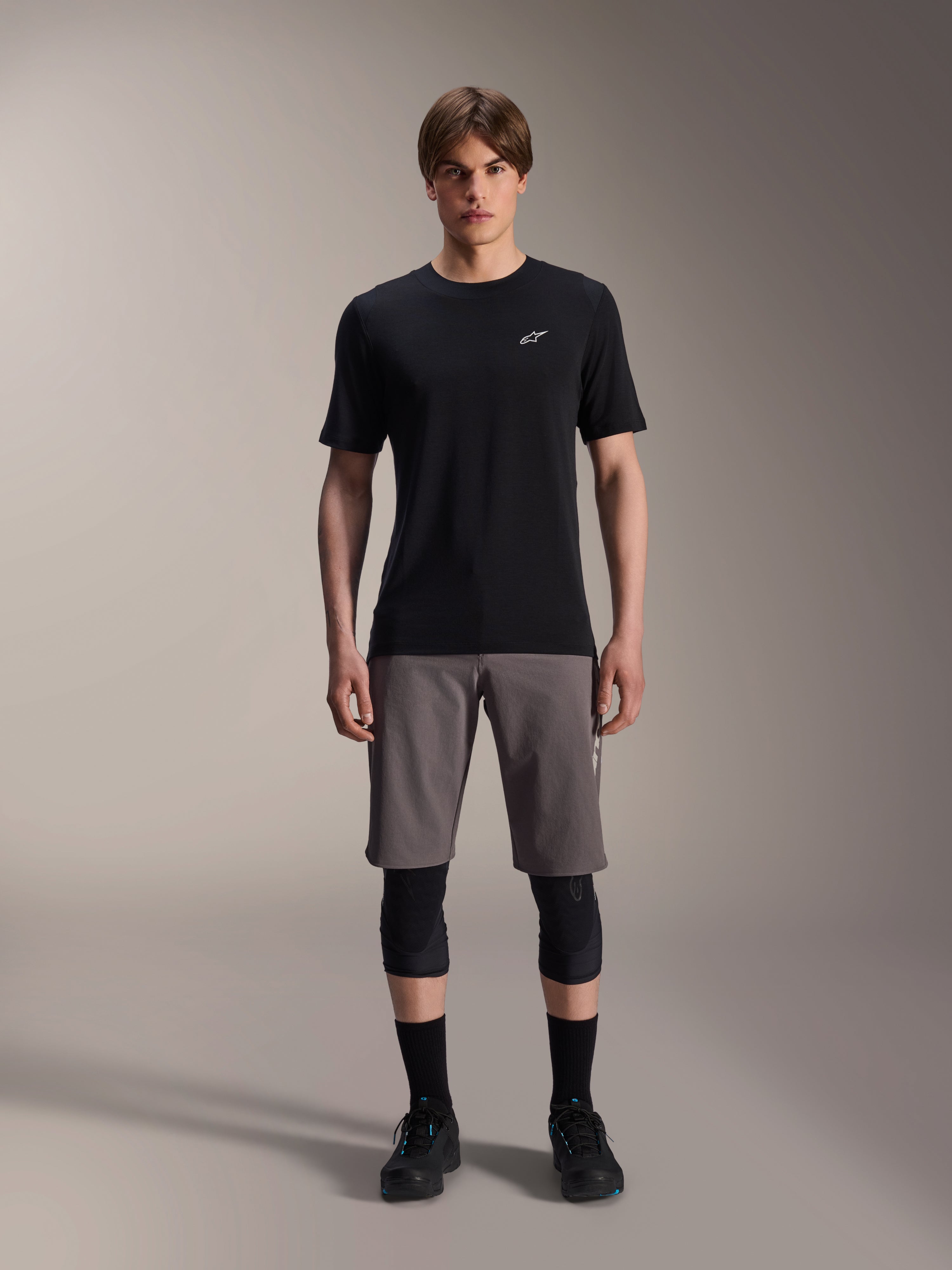 Jersey de manga corta Alpinestars A-Dura Dri Wool, negro, usado por un modelo con pantalones cortos de MTB grises, rodilleras negras y zapatillas de ciclismo negras, con un diseño minimalista con un pequeño logotipo Astars blanco en el pecho, ropa técnica de alto rendimiento para trail.