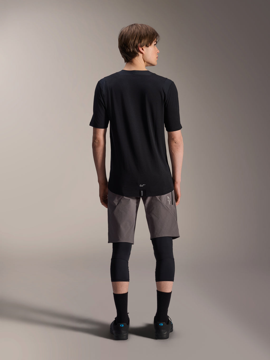 Alpinestars A-Dura Elite Shorts, pantalones cortos de mountain bike en gris oscuro, vista trasera, combinados con un jersey negro de manga corta, rodilleras y calcetines negros, equipamiento técnico de ciclismo para recorridos off-road