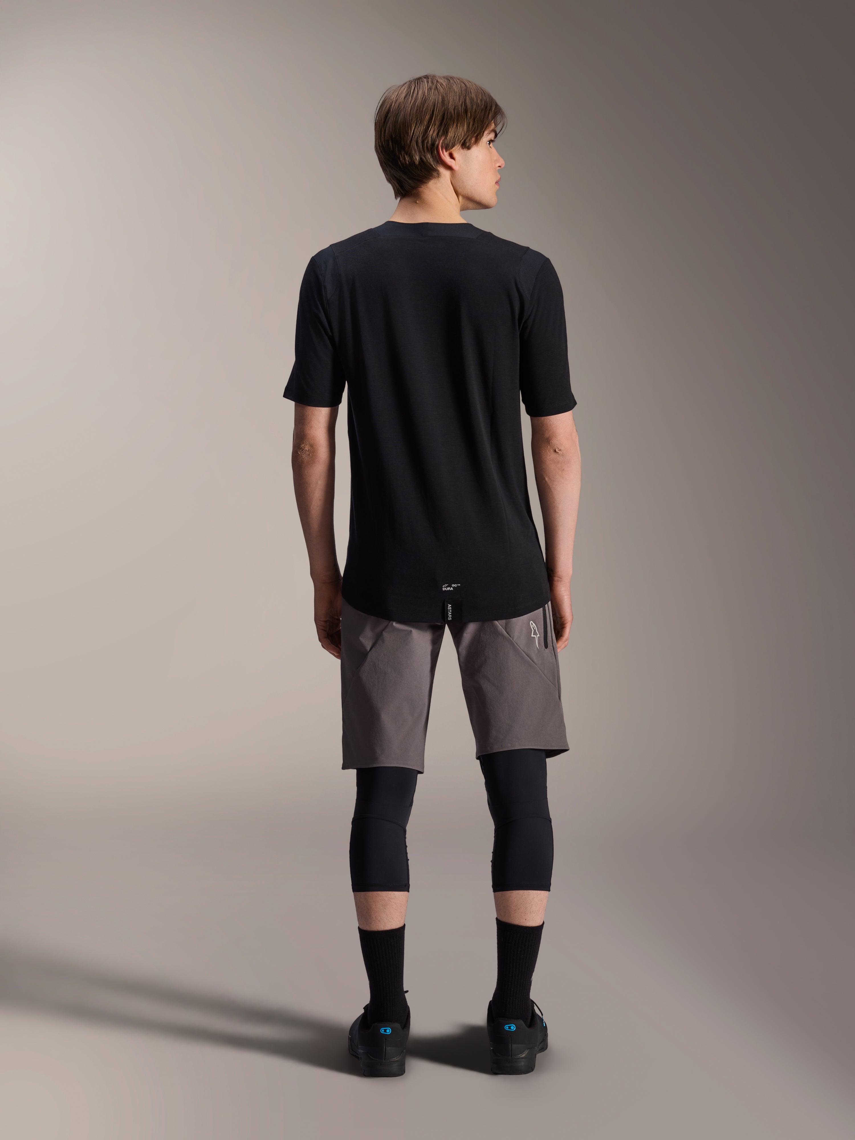 Jersey de manga corta Alpinestars A-Dura Dri Wool, negro, vista trasera usada por un modelo con pantalones cortos de MTB grises y rodilleras negras, con cola trasera más larga y ajuste atlético para ciclismo de montaña