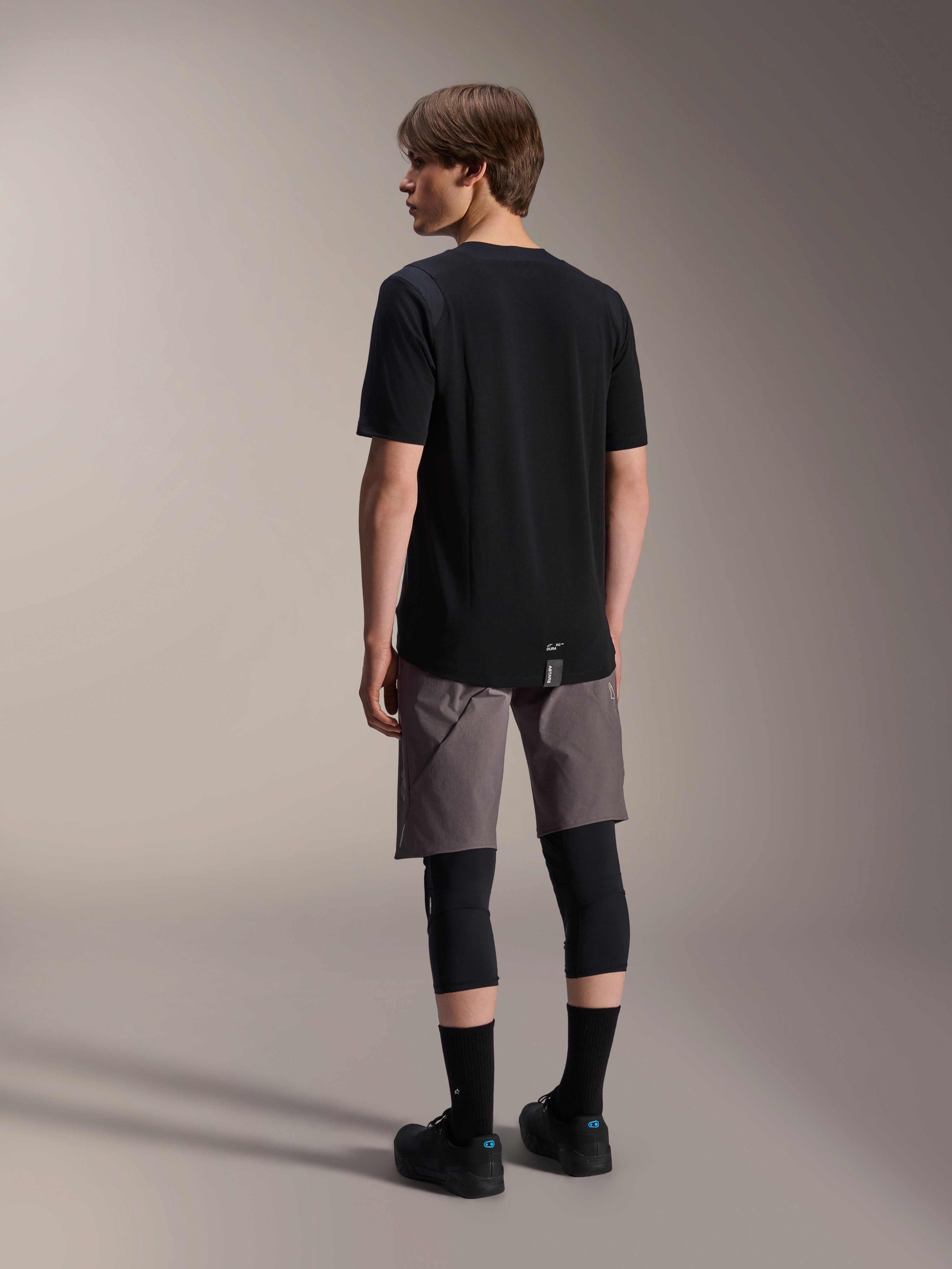 Alpinestars A-Dura Dri Wool Short Sleeve Jersey, negro, vista trasera en modelo, usado con pantalones cortos de ciclismo de montaña grises y rodilleras negras, con dobladillo trasero más largo y un logotipo sutil, ropa técnica de MTB