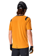Alpinestars A-Dura Dri Wool Jersey de manga corta, oro oscuro, vista trasera de un hombre usando el jersey de manga corta con paneles de malla negra en los hombros, combinado con un casco de MTB negro y guantes, equipamiento de ciclismo de montaña de ajuste regular.
