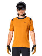 Camiseta de manga corta Alpinestars A-Dura Dri Wool, color oro oscuro, usada con casco de MTB negro, gafas de sol y guantes, con cuello y paneles en los hombros negros, mezcla de lana drirelease transpirable para ciclismo de montaña