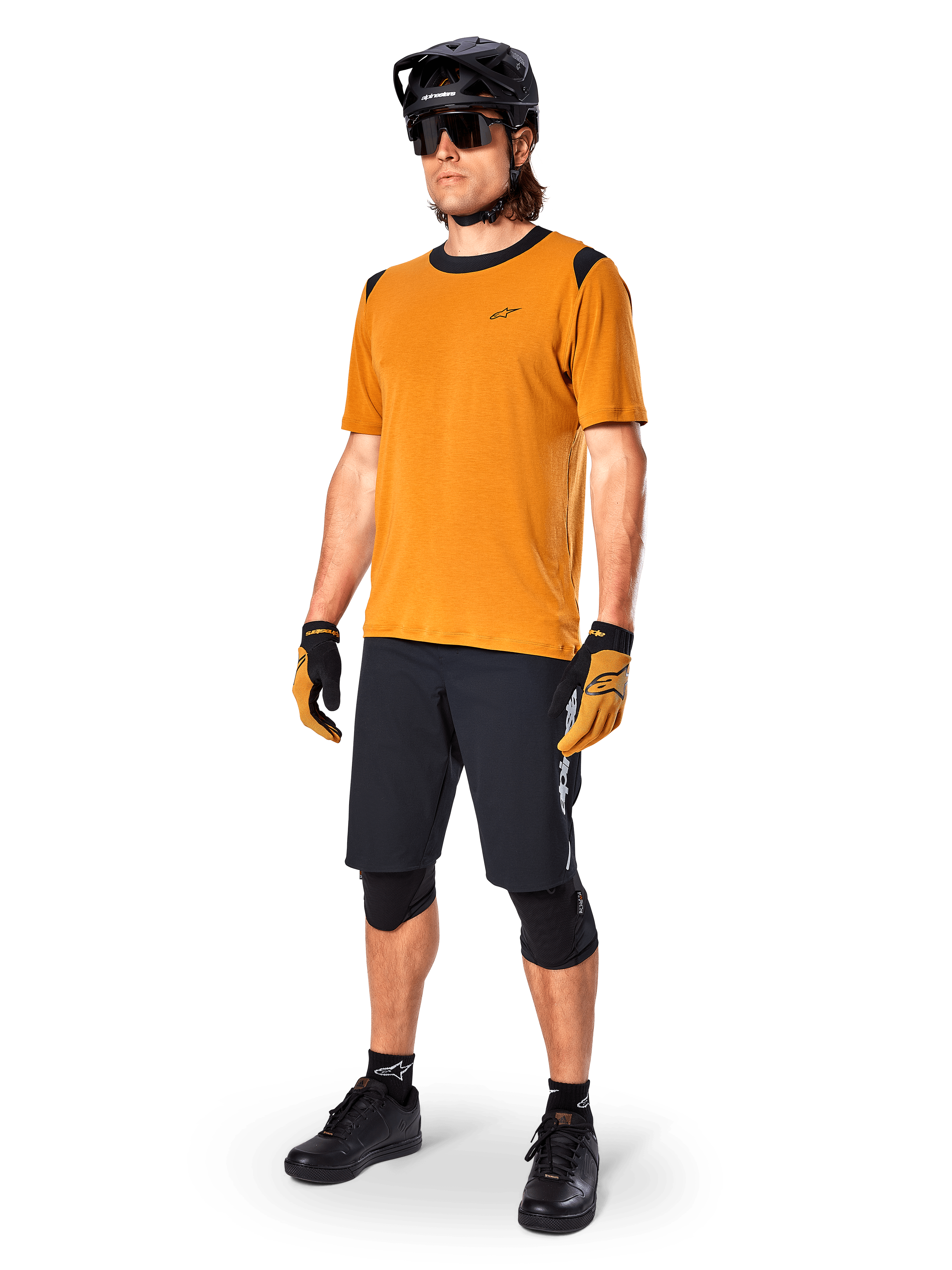 Modelo masculino vistiendo el Jersey Alpinestars A-Dura Dri Wool - Manga Corta en color Dorado Oscuro, jersey de ciclismo de montaña con detalles negros en hombros y cuello, combinado con pantalones cortos de MTB negros, casco negro y gafas de sol, equipamiento técnico de ciclismo todoterreno
