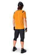 Alpinestars A-Dura Dri Wool Short Sleeve Jersey, jersey de ciclismo, oro oscuro, vista trasera de ciclista de montaña con jersey amarillo mostaza y acentos de malla negra en los hombros, combinado con pantalones cortos negros, rodilleras, casco negro y guantes, equipamiento técnico de ciclismo todoterreno