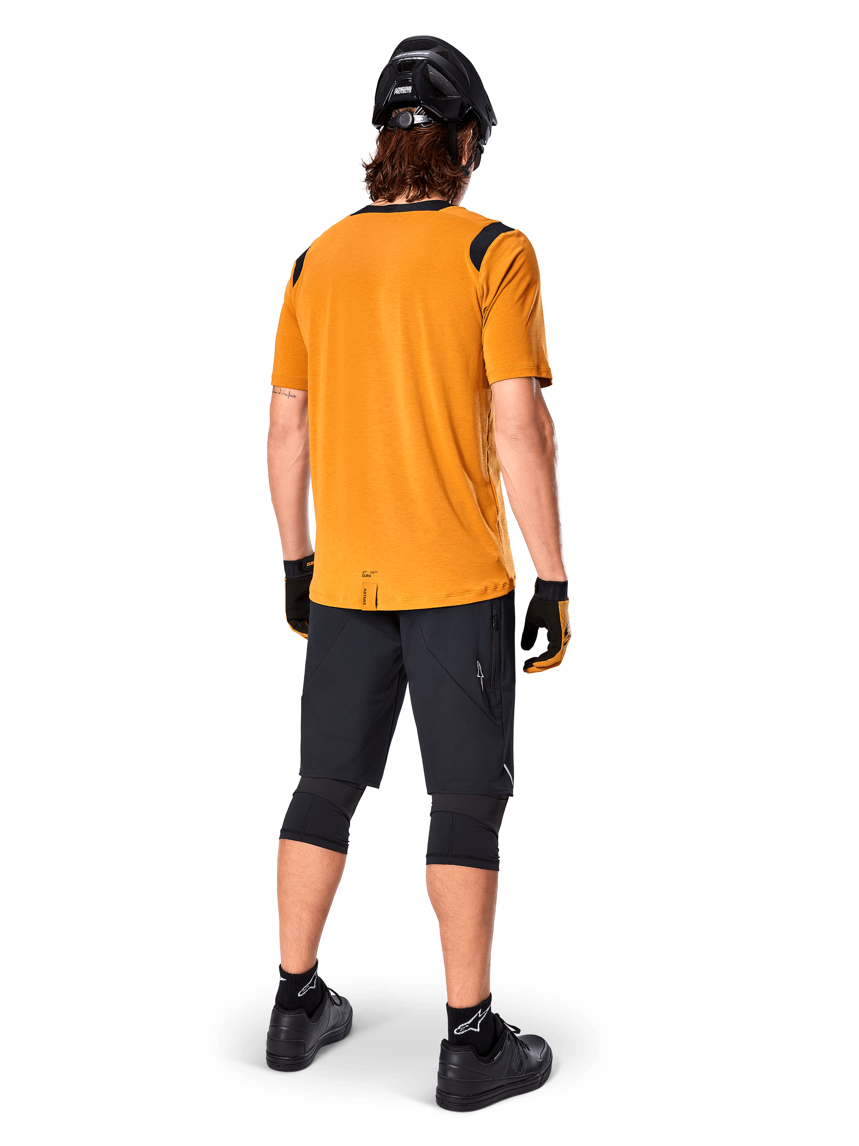 Alpinestars A-Dura Dri Wool Short Sleeve Jersey, jersey de ciclismo, oro oscuro, vista trasera de ciclista de montaña con jersey amarillo mostaza y acentos de malla negra en los hombros, combinado con pantalones cortos negros, rodilleras, casco negro y guantes, equipamiento técnico de ciclismo todoterreno