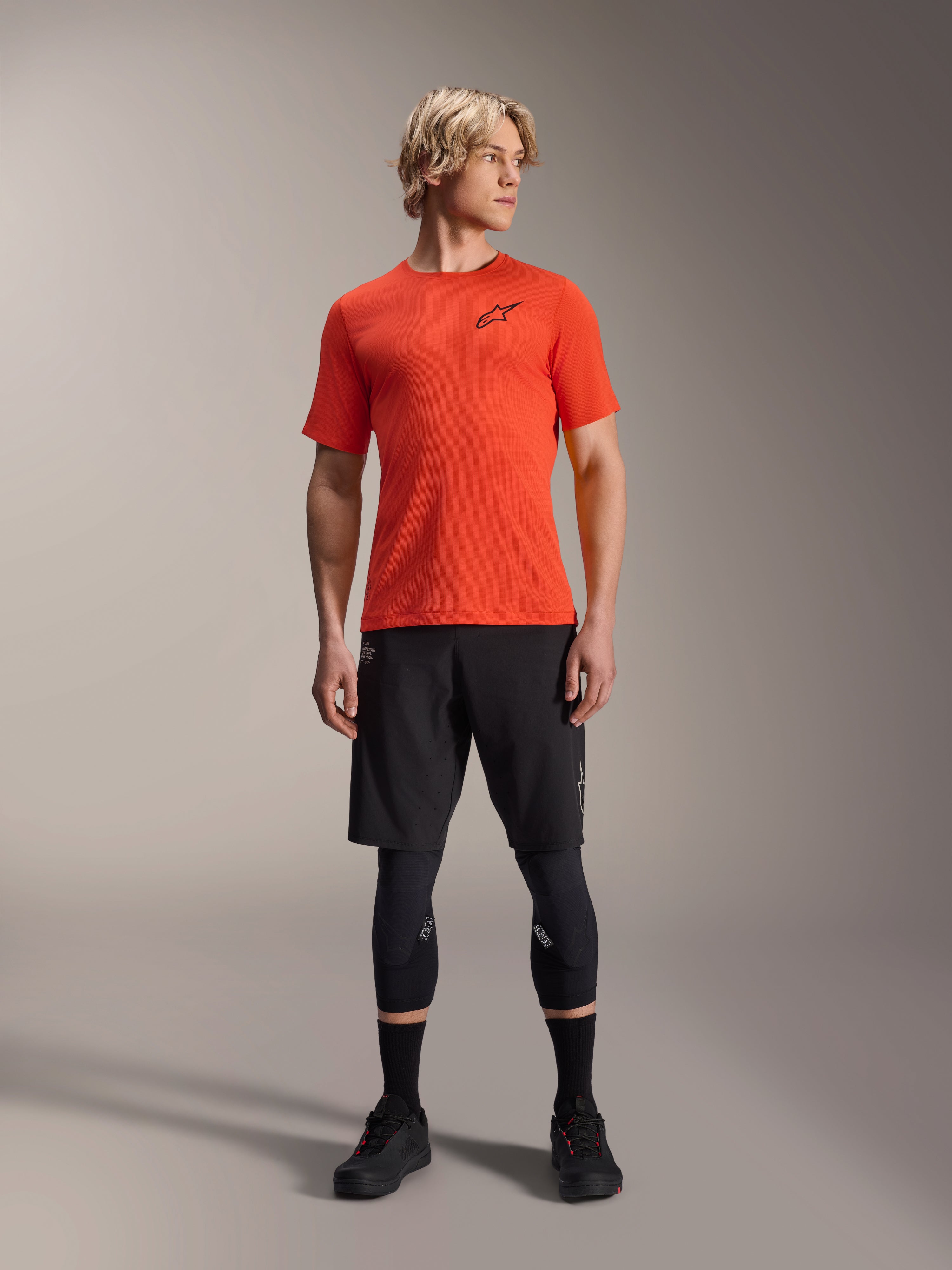 Alpinestars A-Aria Astar Jersey de manga corta, Spicy Orange, usado por un modelo con pantalones cortos de MTB negros y rodilleras, ropa de ciclismo de rendimiento ligera y transpirable para MTB