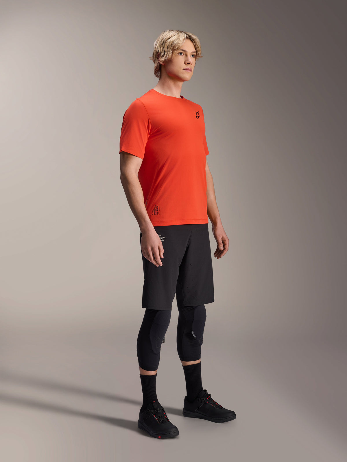 Camiseta de manga corta Alpinestars A-Aria Astar en Spicy Orange, usada por un modelo con pantalones cortos de MTB negros y protecciones de rodilla, ropa de ciclismo de montaña ligera y transpirable, color naranja vibrante con logotipos mínimos en negro