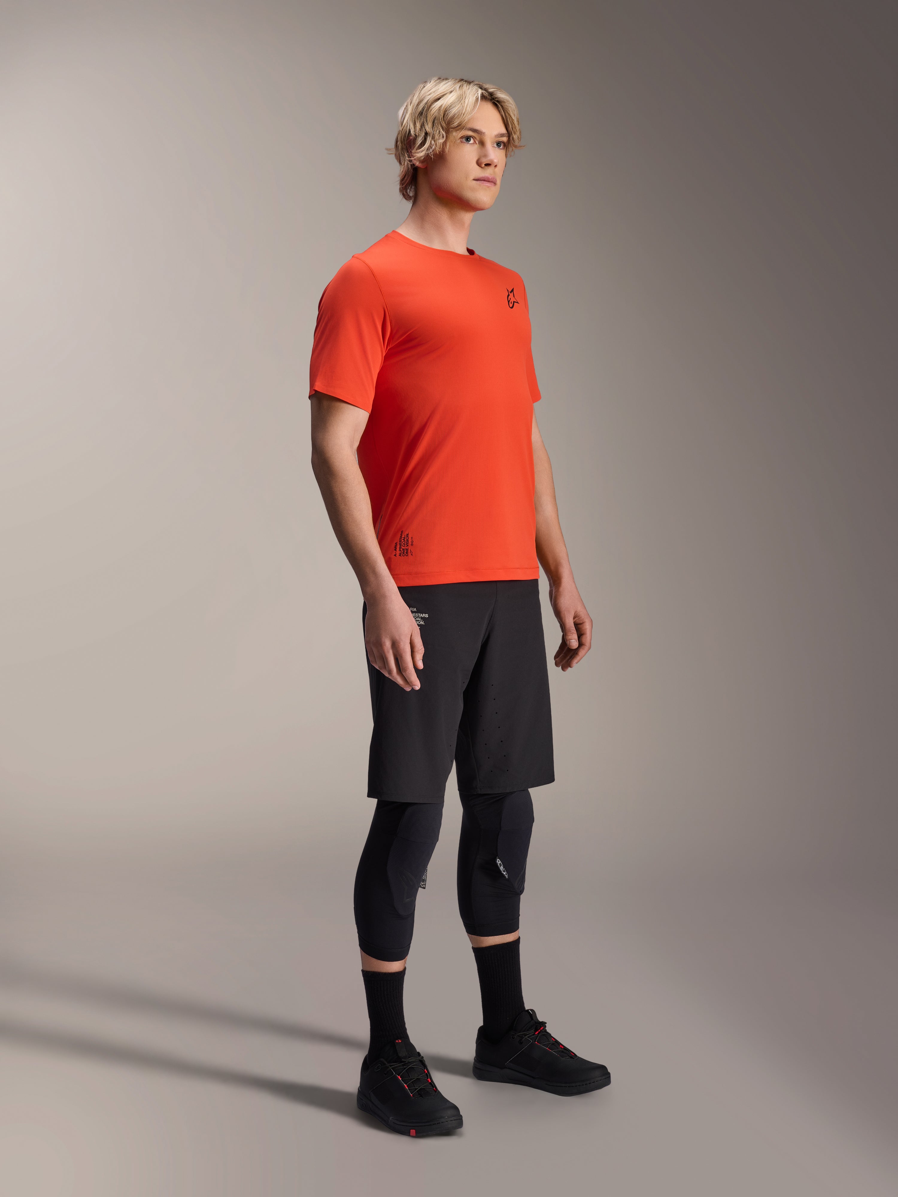 Camiseta de manga corta Alpinestars A-Aria Astar en Spicy Orange, usada por un modelo con pantalones cortos de MTB negros y protecciones de rodilla, ropa de ciclismo de montaña ligera y transpirable, color naranja vibrante con logotipos mínimos en negro