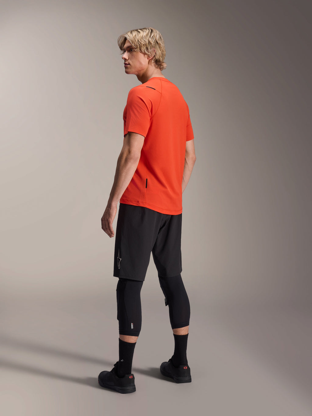 Jersey de manga corta Alpinestars A-Aria Astar, Spicy Orange, vista trasera de tres cuartos del modelo mirando sobre el hombro, combinado con pantalones cortos de MTB negros y rodilleras, ropa técnica para bicicleta de montaña