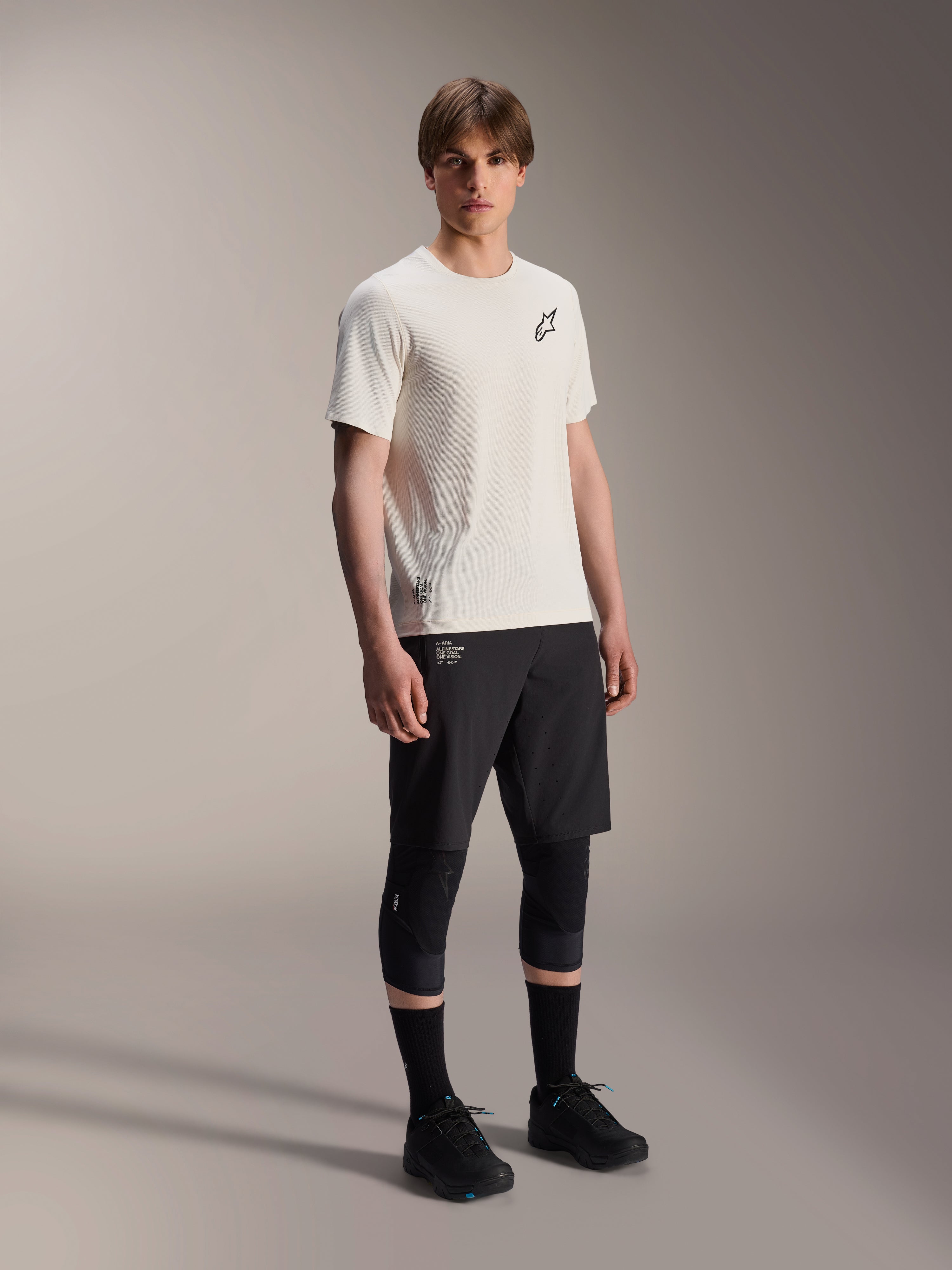 Jersey Alpinestars A-Aria Astar de manga corta, color crudo, con logotipo negro en el pecho, combinado con pantalones cortos de ciclismo y rodilleras negros, equipamiento ligero de alto rendimiento para MTB
