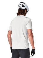 Alpinestars A-Aria Astar Jersey, manga corta, blanco (White Dove), vista trasera de este jersey de ciclismo de montaña ligero y transpirable, usado con casco blanco y guantes de ciclismo negros, con una parte trasera más larga para mayor cobertura