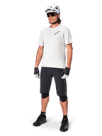 Ciclista de MTB con el jersey Alpinestars A-Aria Astar de manga corta en color White Dove, combinado con pantalones cortos de montaña negros, casco blanco y rodilleras negras, con un pequeño logotipo negro en el pecho, equipamiento de ciclismo coordinado para el rendimiento en senderos