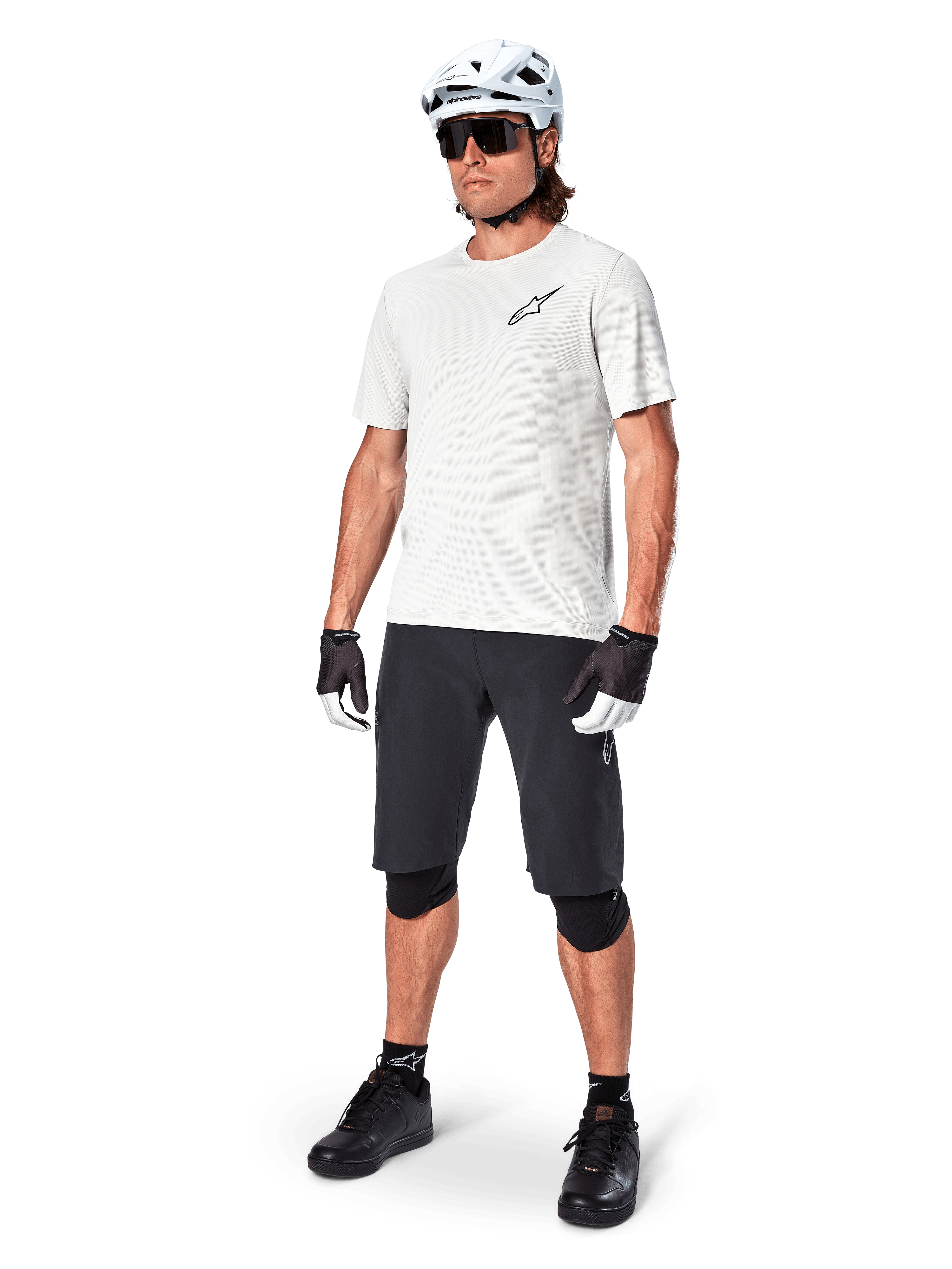 Ciclista de MTB con el jersey Alpinestars A-Aria Astar de manga corta en color White Dove, combinado con pantalones cortos de montaña negros, casco blanco y rodilleras negras, con un pequeño logotipo negro en el pecho, equipamiento de ciclismo coordinado para el rendimiento en senderos