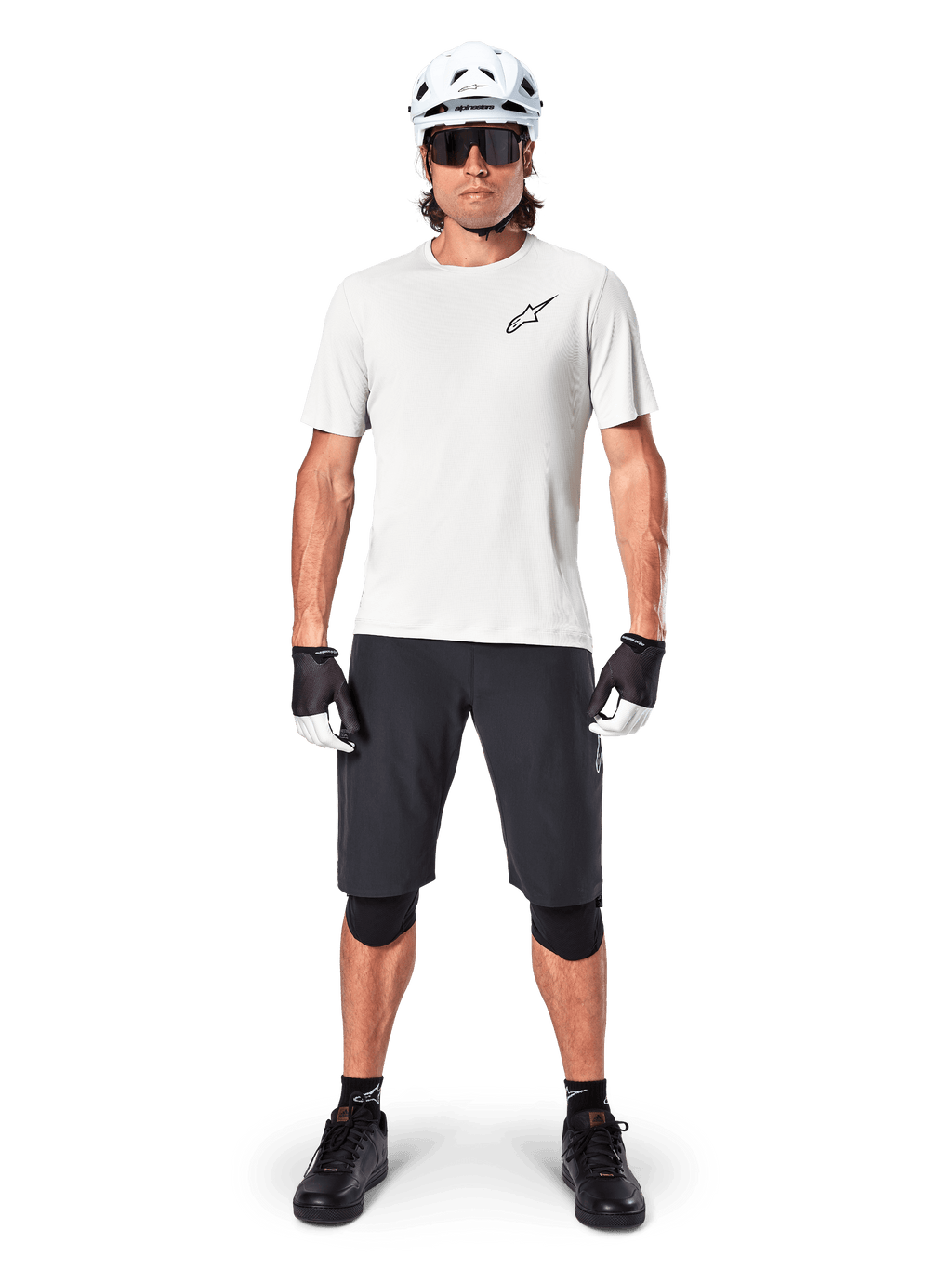 Ciclista masculino con jersey de manga corta Alpinestars A-Aria Astar en color White Dove, con logo Astars negro en el pecho, combinado con pantalones cortos de ciclismo de montaña negros, casco blanco, gafas de sol, rodilleras y guantes, equipamiento completo de ciclismo.