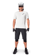 Ciclista masculino con jersey de manga corta Alpinestars A-Aria Astar en color White Dove, con logo Astars negro en el pecho, combinado con pantalones cortos de ciclismo de montaña negros, casco blanco, gafas de sol, rodilleras y guantes, equipamiento completo de ciclismo.