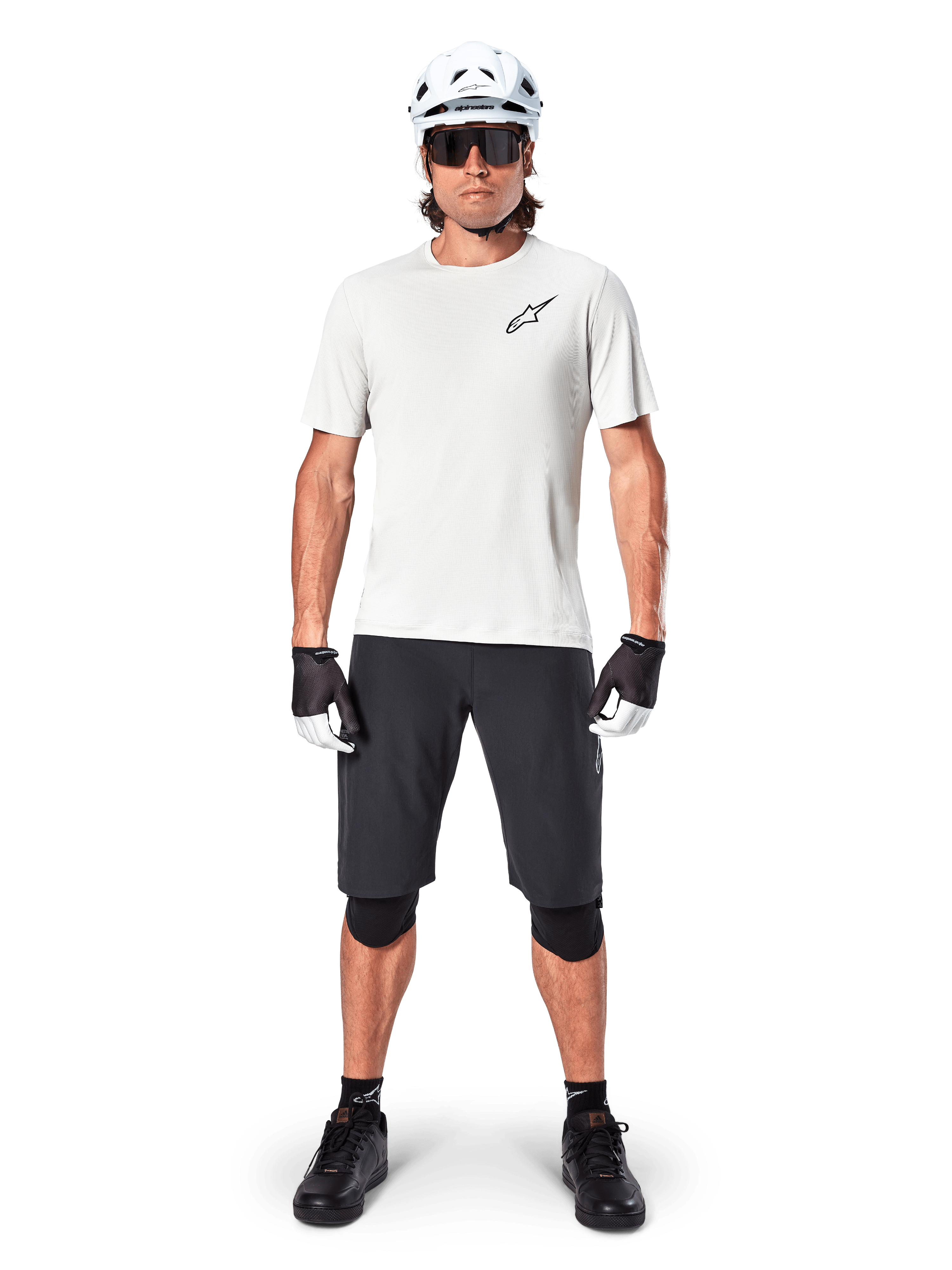 Ciclista masculino con jersey de manga corta Alpinestars A-Aria Astar en color White Dove, con logo Astars negro en el pecho, combinado con pantalones cortos de ciclismo de montaña negros, casco blanco, gafas de sol, rodilleras y guantes, equipamiento completo de ciclismo.
