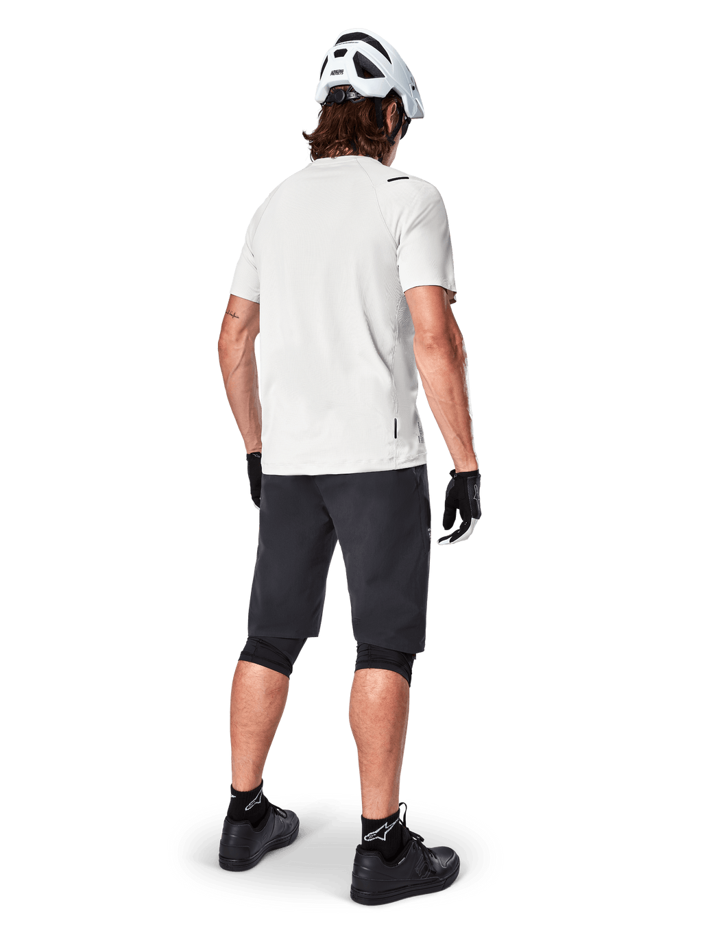 Ciclista con el jersey de manga corta Alpinestars A-Aria Astar en color White Dove, vista trasera, combinado con pantalones cortos de MTB negros, rodilleras y casco blanco, equipamiento ligero y transpirable para ciclismo de montaña todoterreno