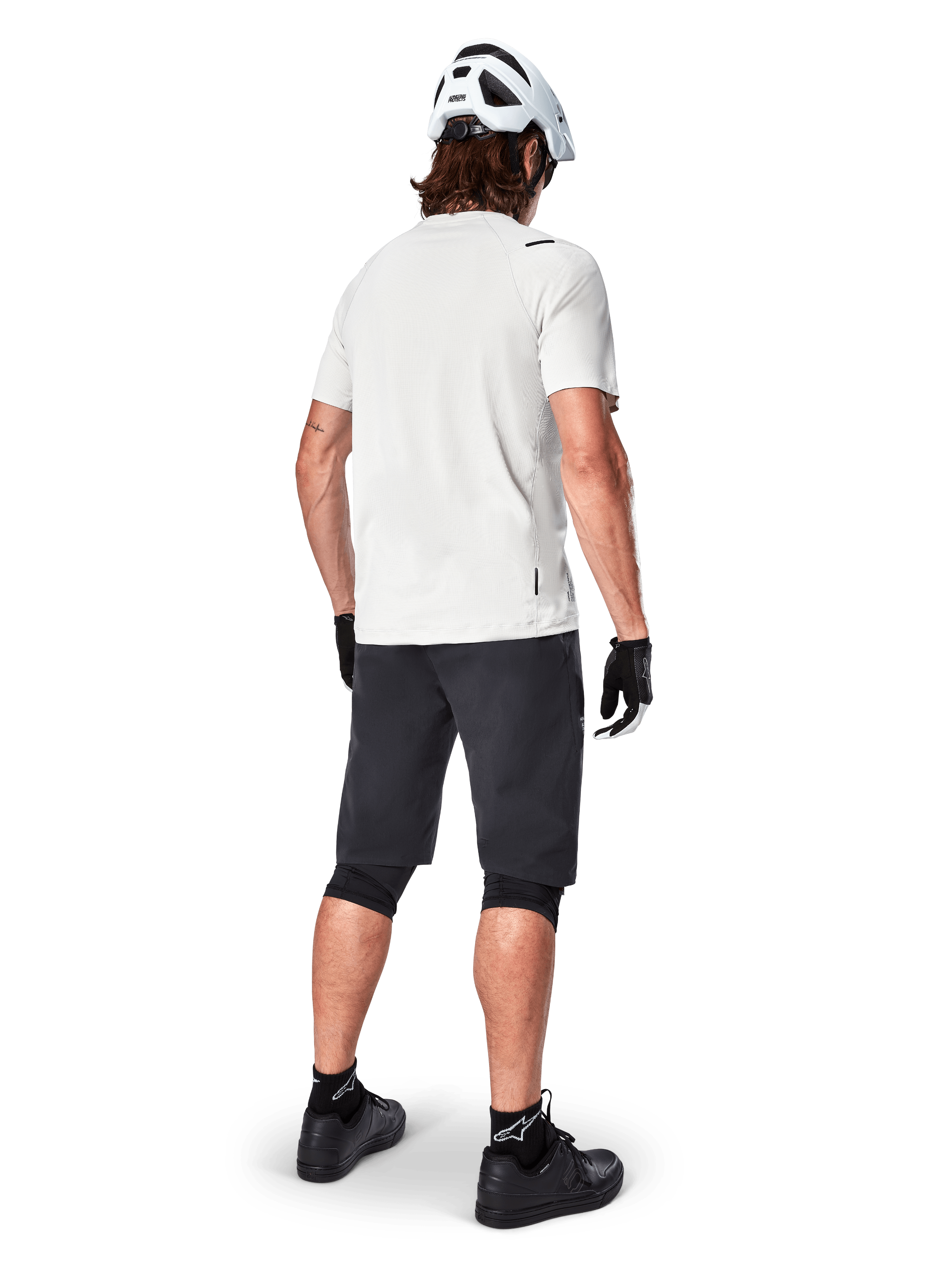 Ciclista con el jersey de manga corta Alpinestars A-Aria Astar en color White Dove, vista trasera, combinado con pantalones cortos de MTB negros, rodilleras y casco blanco, equipamiento ligero y transpirable para ciclismo de montaña todoterreno