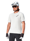 Jersey de manga corta Alpinestars A-Aria Astar, White Dove, usado por un ciclista con casco blanco y gafas de sol oscuras, con un pequeño logotipo de estrella negro en el pecho, equipamiento de alto rendimiento transpirable para ciclismo de montaña