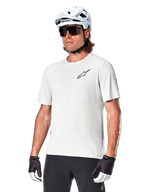 Jersey de manga corta Alpinestars A-Aria Astar, White Dove, usado por un ciclista con casco blanco y gafas de sol oscuras, con un pequeño logotipo de estrella negro en el pecho, equipamiento de alto rendimiento transpirable para ciclismo de montaña