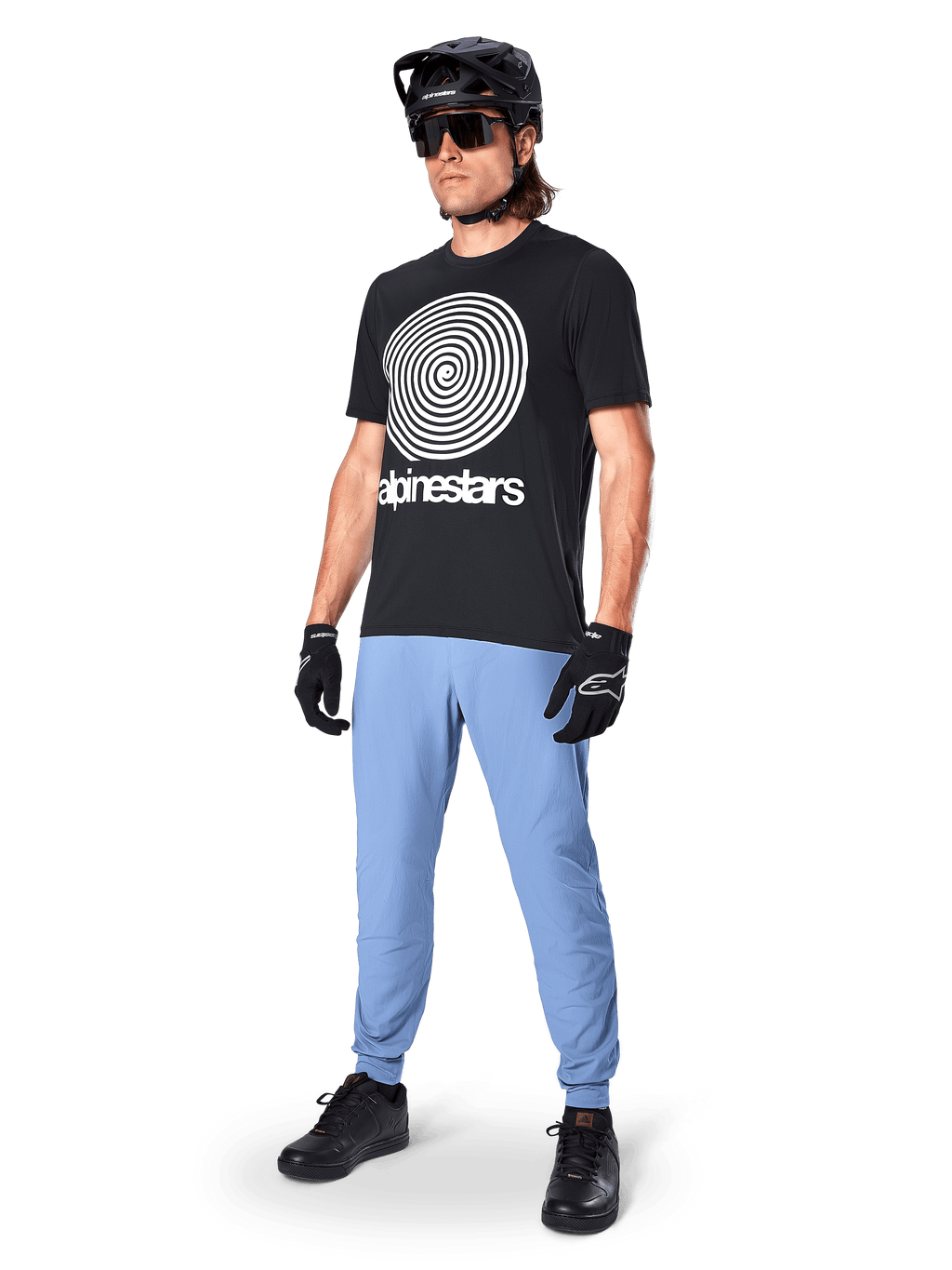 Alpinestars Jersey de Manga Corta A-Dura Oscar, jersey de MTB negro con un gran gráfico en espiral blanco, usado por un ciclista con casco negro, gafas de sol, guantes negros y pantalones azul claro, equipamiento de alto rendimiento para mountain bike
