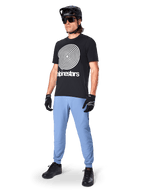 Alpinestars Jersey de Manga Corta A-Dura Oscar, jersey de MTB negro con un gran gráfico en espiral blanco, usado por un ciclista con casco negro, gafas de sol, guantes negros y pantalones azul claro, equipamiento de alto rendimiento para mountain bike