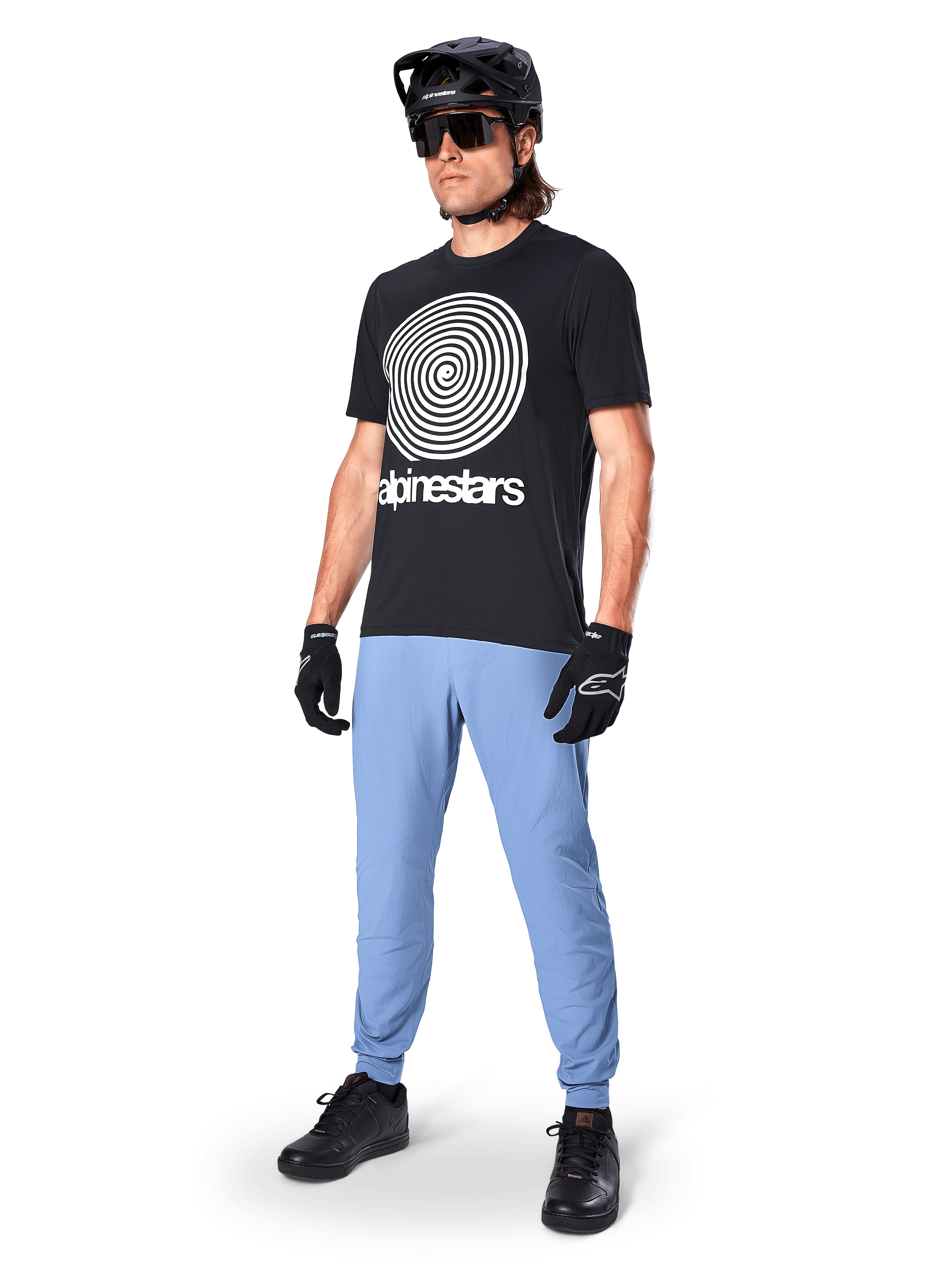 Alpinestars Jersey de Manga Corta A-Dura Oscar, jersey de MTB negro con un gran gráfico en espiral blanco, usado por un ciclista con casco negro, gafas de sol, guantes negros y pantalones azul claro, equipamiento de alto rendimiento para mountain bike